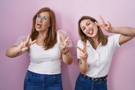 Photo for Hispanic mother and daughter wearing casual white t shirt over pink background smiling with tongue out showing fingers of both hands doing victory sign. number two. - Royalty Free Image