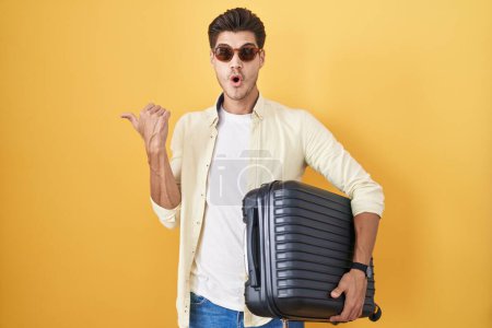 Foto de Joven hombre hispano sosteniendo maleta yendo de vacaciones de verano sorprendido señalando con el dedo de la mano hacia un lado, boca abierta expresión asombrada. - Imagen libre de derechos