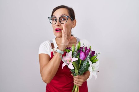 Foto de Mujer morena de mediana edad usando delantal trabajando en floristería sosteniendo ramo de mano en boca contando rumores secretos, susurrando conversación maliciosa - Imagen libre de derechos