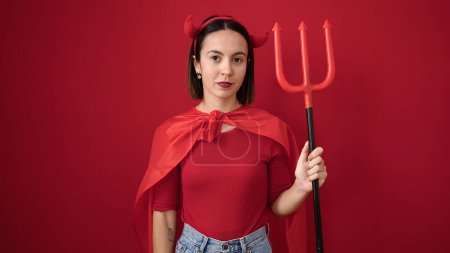 Foto de Joven hermosa mujer hispana con traje de diablo sosteniendo tridente sobre fondo rojo aislado - Imagen libre de derechos