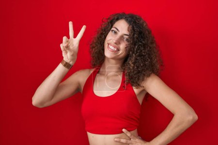 Téléchargez les photos : Femme hispanique avec les cheveux bouclés debout sur fond rouge souriant en regardant la caméra montrant les doigts faisant signe de victoire. numéro deux. - en image libre de droit