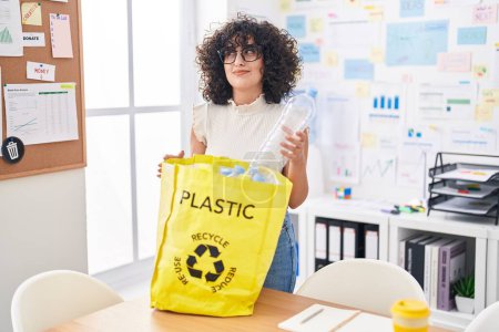 Foto de Joven mujer de Oriente Medio sosteniendo bolsa de reciclaje con botellas de plástico en la oficina sonriendo mirando hacia un lado y mirando hacia otro pensando. - Imagen libre de derechos