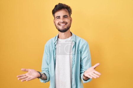 Foto de Joven hombre hispano con tatuajes de pie sobre fondo amarillo sonriente alegre con los brazos abiertos como bienvenida amistosa, saludos positivos y confiados - Imagen libre de derechos
