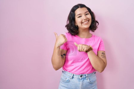 Téléchargez les photos : Young hispanic woman standing over pink background pointing to the back behind with hand and thumbs up, smiling confident - en image libre de droit
