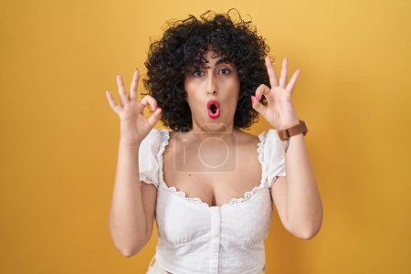 Photo for Young brunette woman with curly hair standing over yellow background looking surprised and shocked doing ok approval symbol with fingers. crazy expression - Royalty Free Image