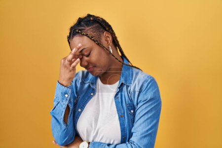 Photo for African american woman with braids standing over yellow background tired rubbing nose and eyes feeling fatigue and headache. stress and frustration concept. - Royalty Free Image