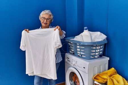 Téléchargez les photos : Senior femme aux cheveux gris souriant confiant tenant t-shirt blanc à la buanderie - en image libre de droit