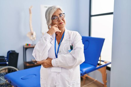 Photo for Middle age woman with grey hair working at pain recovery clinic looking confident at the camera smiling with crossed arms and hand raised on chin. thinking positive. - Royalty Free Image