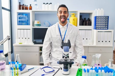 Foto de Un joven hispano con barba trabajando en el laboratorio científico sacando la lengua feliz con expresión divertida. concepto de emoción. - Imagen libre de derechos