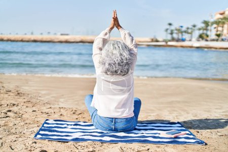 Téléchargez les photos : Femme d'âge moyen faisant de l'exercice de yoga assis sur une serviette au bord de la mer - en image libre de droit