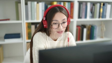 Téléchargez les photos : Jeune belle étudiante hispanique souriante confiante ayant un appel vidéo à la bibliothèque de l'université - en image libre de droit