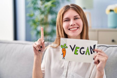 Foto de Hermosa mujer sosteniendo pancarta con palabra vegana sonriendo con una idea o pregunta señalando el dedo con la cara feliz, número uno - Imagen libre de derechos