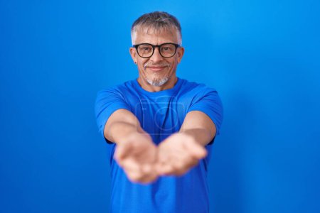 Photo for Hispanic man with grey hair standing over blue background smiling with hands palms together receiving or giving gesture. hold and protection - Royalty Free Image
