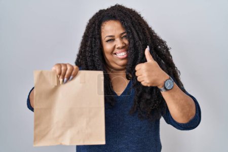 Téléchargez les photos : Plus taille hispanique femme tenant emporter sac en papier souriant heureux et positif, pouce vers le haut faire excellent et signe d'approbation - en image libre de droit