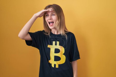 Foto de Mujer rubia caucásica con camiseta bitcoin muy feliz y sonriente mirando lejos con la mano sobre la cabeza. concepto de búsqueda. - Imagen libre de derechos