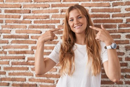 Téléchargez les photos : Jeune femme caucasienne debout sur le mur de briques souriant joyeuse montrant et pointant du doigt les dents et la bouche. concept de santé dentaire. - en image libre de droit