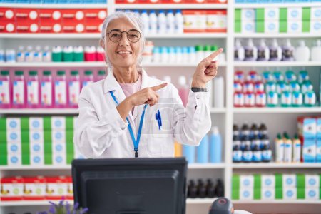 Photo for Middle age woman with tattoos working at pharmacy drugstore smiling and looking at the camera pointing with two hands and fingers to the side. - Royalty Free Image