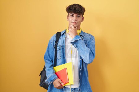 Photo for Hispanic teenager wearing student backpack and holding books looking confident at the camera smiling with crossed arms and hand raised on chin. thinking positive. - Royalty Free Image