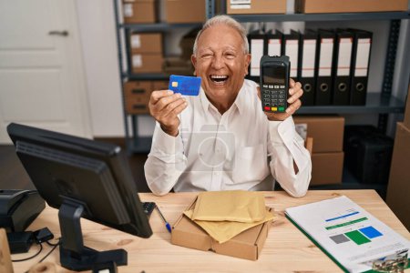 Foto de Hombre mayor con el pelo gris que trabaja en el comercio electrónico de la pequeña empresa sosteniendo la tarjeta de crédito y el datáfono sonriendo y riendo fuerte en voz alta porque broma loca divertida. - Imagen libre de derechos