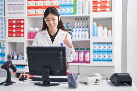 Téléchargez les photos : Jeune femme chinoise travaillant à la pharmacie pharmacie portant casque souriant heureux et positif, pouce levé faire excellent et signe d'approbation - en image libre de droit