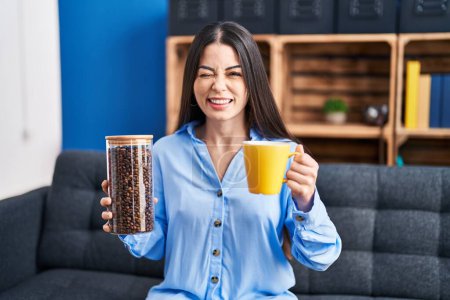 Téléchargez les photos : Jeune femme brune tenant des grains de café et une tasse de café clignant des yeux en regardant la caméra avec expression sexy, visage joyeux et heureux. - en image libre de droit