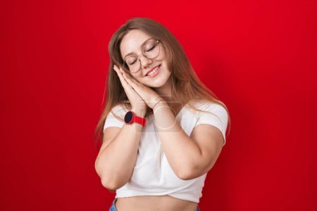Photo for Young caucasian woman standing over red background sleeping tired dreaming and posing with hands together while smiling with closed eyes. - Royalty Free Image