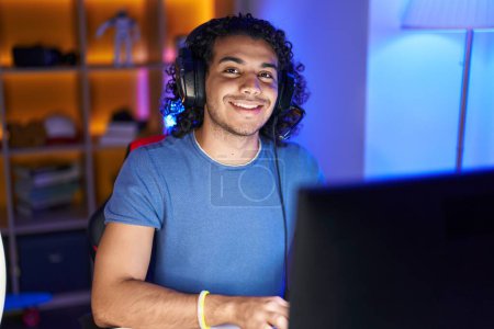 Foto de Joven hombre latino streamer sonriendo confiado sentado en la mesa en la sala de juegos - Imagen libre de derechos