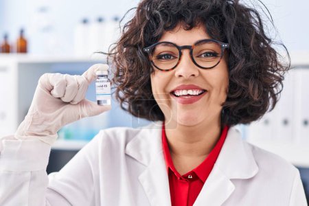 Téléchargez les photos : Médecin hispanique femme aux cheveux bouclés tenant le vaccin à l'air positif et heureux debout et souriant avec un sourire confiant montrant les dents - en image libre de droit