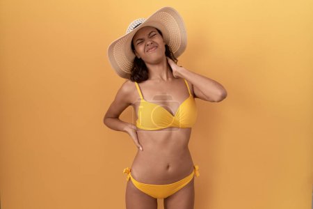 Téléchargez les photos : Jeune femme hispanique portant un bikini et un chapeau d'été souffrant de douleurs au cou, touchant le cou avec la main, douleurs musculaires - en image libre de droit
