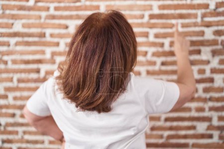 Photo for Senior woman with glasses standing over bricks wall posing backwards pointing ahead with finger hand - Royalty Free Image