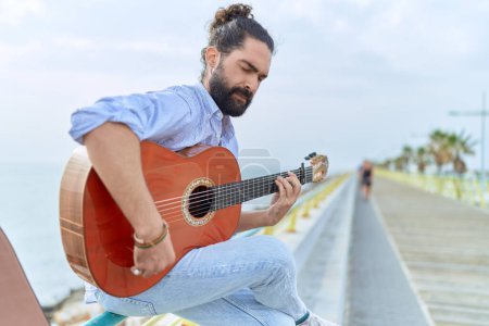 Téléchargez les photos : Young hispanic man musician playing classical guitar sitting on balustrade at seaside - en image libre de droit