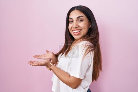 Téléchargez les photos : Jeune femme arabe debout sur fond rose pointant vers le côté avec les mains ouvertes paumes montrant l'espace de copie, présentant la publicité souriant heureux excité - en image libre de droit