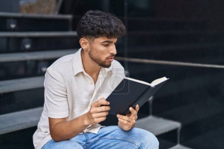 Téléchargez les photos : Jeune homme arabe lecture livre assis sur les escaliers à la rue - en image libre de droit