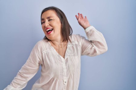 Foto de Mujer hispana de mediana edad de pie sobre fondo azul bailando feliz y alegre, sonriente movimiento casual y seguro escuchando música - Imagen libre de derechos