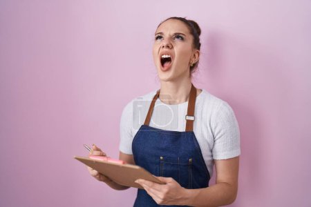Photo for Young hispanic girl wearing professional waitress apron taking order angry and mad screaming frustrated and furious, shouting with anger looking up. - Royalty Free Image