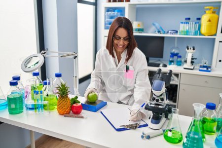 Photo for Young beautiful hispanic woman scientist weighing apple writing on document at laboratory - Royalty Free Image