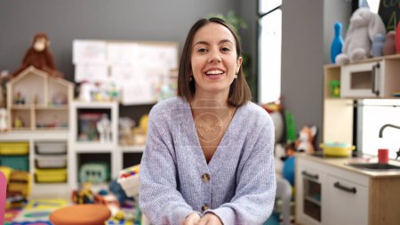 Photo for Young beautiful hispanic woman teacher smiling confident sitting on table at kindergarten - Royalty Free Image