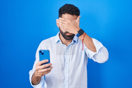 Photo for Hispanic man with beard using smartphone typing message covering eyes with hand, looking serious and sad. sightless, hiding and rejection concept - Royalty Free Image