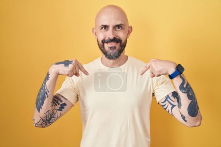 Foto de Hombre hispano con tatuajes de pie sobre fondo amarillo mirando confiado con sonrisa en la cara, señalándose con los dedos orgullosos y felices. - Imagen libre de derechos