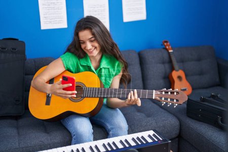 Téléchargez les photos : Jeune musicienne hispanique jouant de la guitare classique en utilisant un smartphone au studio de musique - en image libre de droit
