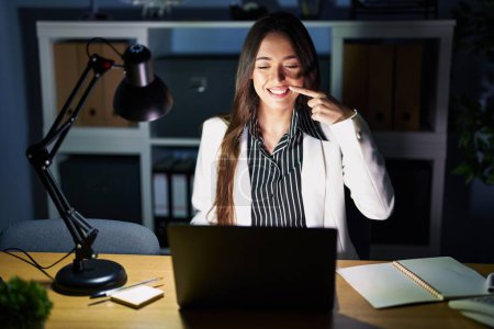 Photo for Young brunette woman working at the office at night with laptop pointing with hand finger to face and nose, smiling cheerful. beauty concept - Royalty Free Image
