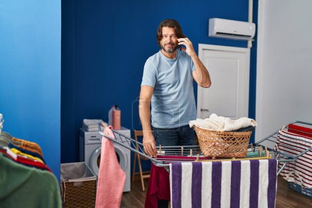 Photo for Middle age man talking on smartphone hanging clothes on clothesline at laundry room - Royalty Free Image