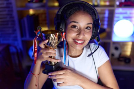 Photo for Young arab woman playing video games holding trophy looking positive and happy standing and smiling with a confident smile showing teeth - Royalty Free Image