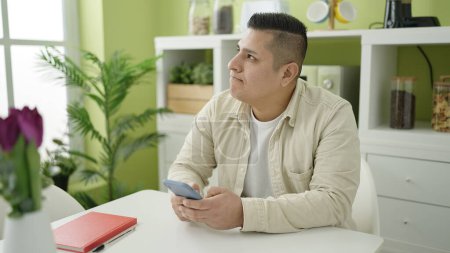 Foto de Joven hombre hispano usando smartphone sentado en la mesa en el comedor - Imagen libre de derechos