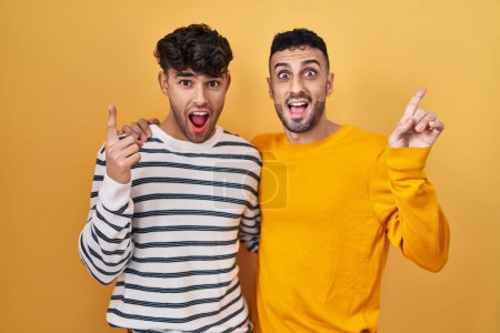Téléchargez les photos : Jeune couple gay hispanique debout sur fond jaune souriant étonné et surpris et pointant vers le haut avec les doigts et les bras levés. - en image libre de droit