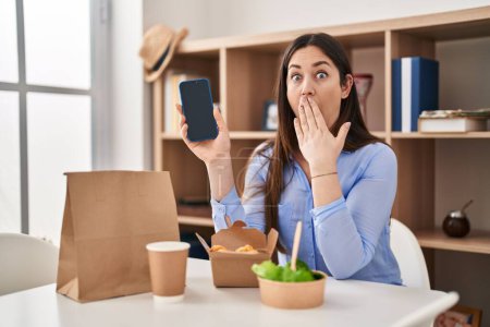 Photo for Young brunette woman eating take away food at home showing smartphone screen covering mouth with hand, shocked and afraid for mistake. surprised expression - Royalty Free Image