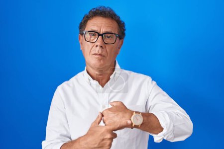 Photo for Middle age hispanic man standing over blue background in hurry pointing to watch time, impatience, looking at the camera with relaxed expression - Royalty Free Image