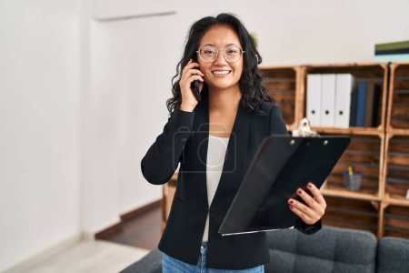 Photo for Young chinese woman psychologist holding clipboard talking on the smartphone at clinic - Royalty Free Image