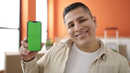 Foto de Joven hombre hispano sonriendo confiado mostrando la pantalla de teléfono inteligente en un nuevo hogar - Imagen libre de derechos