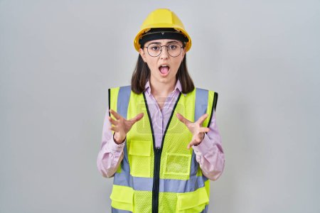 Photo for Hispanic girl wearing builder uniform and hardhat crazy and mad shouting and yelling with aggressive expression and arms raised. frustration concept. - Royalty Free Image
